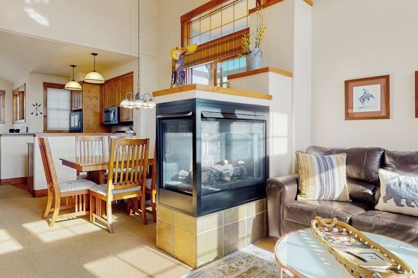 A modern kitchen with a central island, a dining area with wooden chairs, a fireplace, and a living room with a leather couch and a coffee table. - Ski-in/out condo with amazing views, washer/dryer, fireplace, & hot tub (Granby)