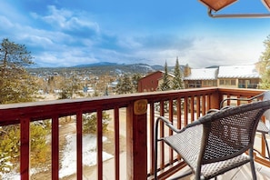Ein Balkon mit Korbstuhl, Holzgeländer und Blick auf eine verschneite Landschaft mit Gebäuden und Bergen in der Ferne.