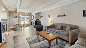 A living room with a sofa, a coffee table, and a TV. There is a dining area with chairs and a table. The room has a wooden floor and a ceiling with exposed beams. - Shuttle to lifts from condo w/ panoramic views (Breckenridge)