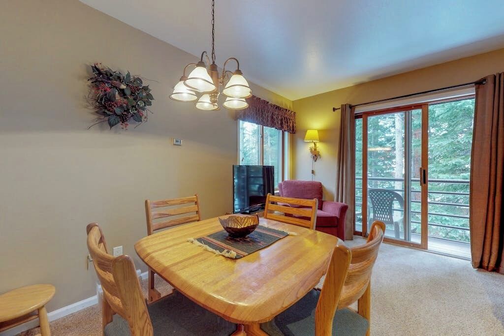 Un coin repas avec une table en bois, des chaises, un lustre, un téléviseur et une vue sur l’extérieur, accessible par une porte-fenêtre coulissante.