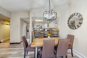 Uma área de refeições com uma mesa e cadeiras de madeira, um candelabro e uma cozinha ao fundo.