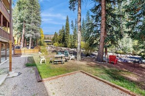 Un espace extérieur aménagé avec un foyer en gravier, une table de pique-nique et des chaises, entouré de grands arbres et d’un bâtiment doté d’une terrasse en bois.