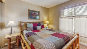 Een slaapkamer met een houten bedframe, een quilt, twee bedlampjes, een ingelijst beeld aan de muur, en een raam met zonwering.