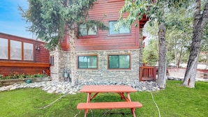 Una casa de dos pisos con fachada de piedra, una terraza de madera y una mesa de picnic en el jardín delantero.