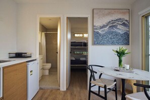A compact kitchen with a dishwasher, a dining area with a table and chairs, a shower, and a bed in the adjacent room. - Clyde B 13 - Family Cabin Sleeps 6 with Bunks (Mollymook)