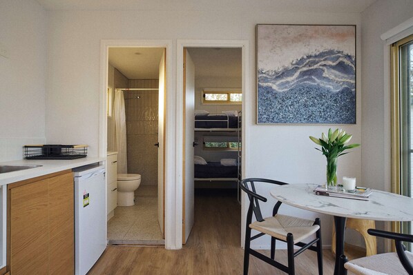 Una cocina compacta con lavavajillas, un comedor con mesa y sillas, una ducha y una cama en la habitación contigua.