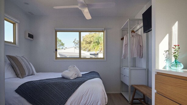 A bedroom with a bed, a window, a ceiling fan, a wardrobe, and a bench. - Clyde B 13 - Family Cabin Sleeps 6 with Bunks (Mollymook)