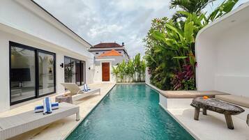 A swimming pool with lounge chairs and a stone bench, surrounded by lush greenery and a white building with large windows.