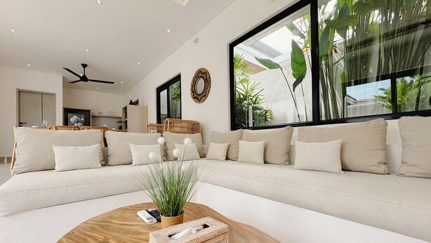 A modern living room with a large sectional sofa, a round wooden coffee table, and large windows with indoor plants. - Canggu Luxury Villa – Private Pool (Badung)