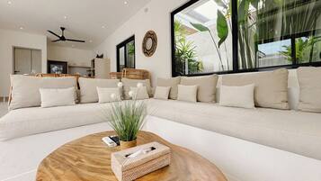 A modern living room with a large sectional sofa, a round wooden coffee table, and large windows with indoor plants.