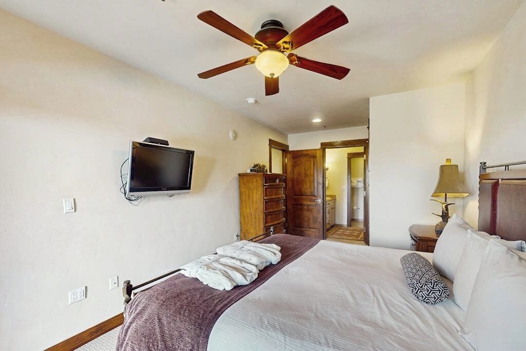 Un dormitorio con cama, ventilador de techo, televisor, cómoda y lámpara.