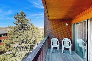 A wooden balcony with two white plastic chairs, a glass door, and a view of trees and buildings. - Spacious, in-town condo with balcony, pools, hot tub, tennis - walk to lifts (Breckenridge)