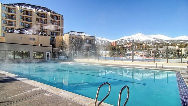 A swimming pool with a view of snow-capped mountains and a multi-story building in the background. - Main Street condo with mountain view, fireplace, pool & hot tubs - walk to lifts (Breckenridge)