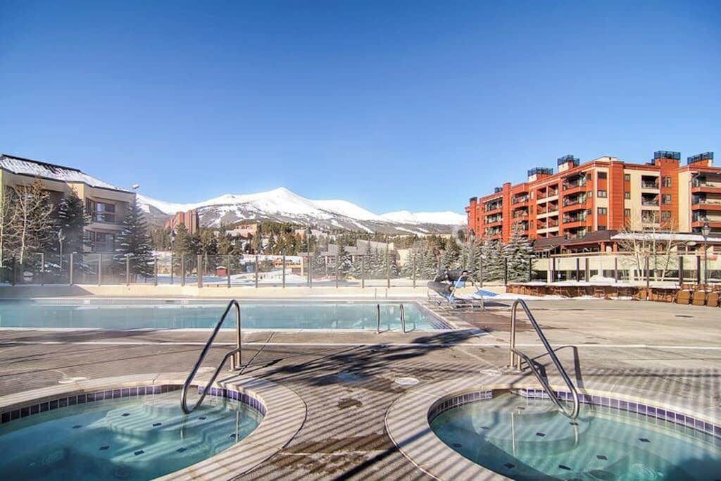 A pool area with hot tubs, a mountain in the background, and a red building with balconies.
