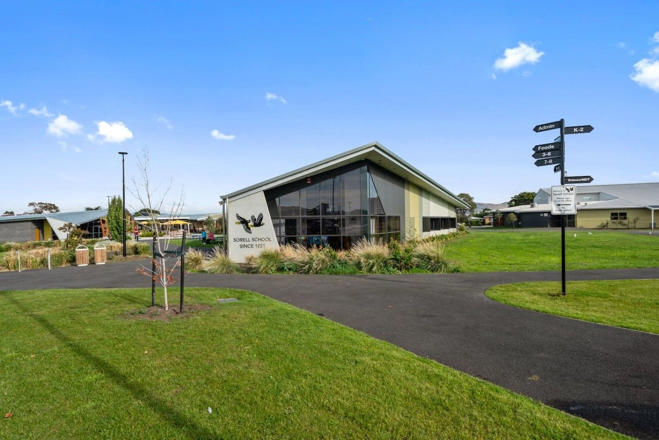A modern school building with a clear sky, a signpost with directions, and a grassy area in front.