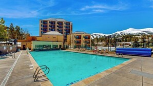 A swimming pool with lounge chairs, a building with multiple floors, and a mountain in the background. - Top-floor downtown condo with pool, hot tubs & gym, walk to ski lifts (Breckenridge)