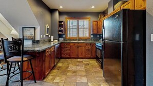 A kitchen with wooden cabinets, a black refrigerator, a window with blinds, and a bar stool. - Top-floor downtown condo with pool, hot tubs & gym, walk to ski lifts (Breckenridge)