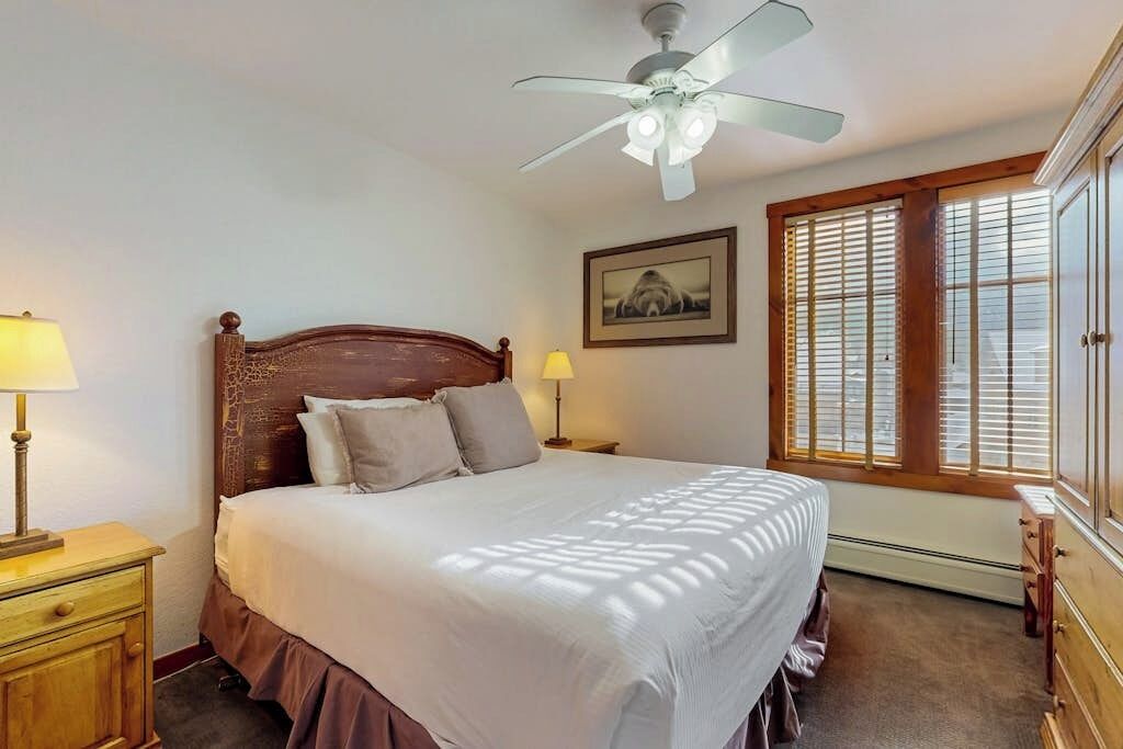 A bedroom with a wooden headboard, a bed with white bedding, a ceiling fan, a window with blinds, a framed picture on the wall, and a nightstand with a lamp.