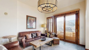 A living room with a brown leather sofa, a wooden coffee table, a round side table, and a large window with a view of mountains. - Mountain condo with pool, hot tubs, gym & steam room - near skiing & hiking (Breckenridge)