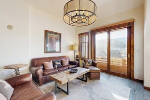 A living room with a brown leather sofa, a wooden coffee table, a round side table, and a large window with a view of mountains. - Mountain condo with pool, hot tubs, gym & steam room - near skiing & hiking (Breckenridge)