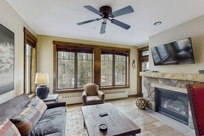 Una sala de estar con chimenea, ventilador de techo, televisor y un amplio ventanal.