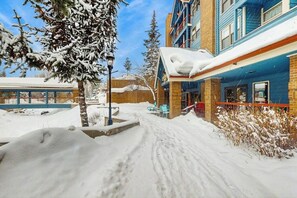 A snow-covered outdoor area with a building featuring a blue and brick facade, a covered seating area, and snow-laden trees. - Beautiful downtown condo with WiFi, pool, hot tub, gym, & balcony - near lifts (Breckenridge)