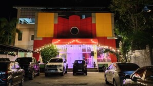 Um edifício com uma entrada ampla, vários carros estacionados e fachada bem iluminada.