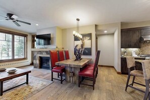 A modern kitchen with a stone fireplace, a dining area with red upholstered chairs, and a large window with blinds. - Modern mountain condo with balcony, pool & hot tubs, walk to lifts/downtown (Breckenridge)