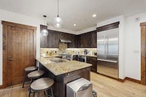 A modern kitchen with dark wood cabinets, stainless steel appliances, and a central island with stools. - Beautiful condo with walk-to-lift location, hot tubs, pool, gym & theater (Breckenridge)