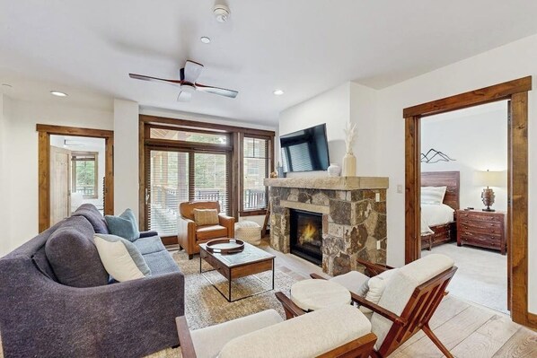 A living room with a stone fireplace, a ceiling fan, and a television mounted on the wall. - Beautiful condo with walk-to-lift location, hot tubs, pool, gym & theater (Breckenridge)