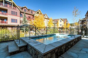 A swimming pool with a stone border, surrounded by a metal fence, and adjacent to multi-story buildings with balconies. - Beautiful condo with walk-to-lift location, hot tubs, pool, gym & theater (Breckenridge)