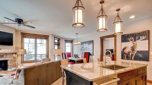 Una cocina con isla central, iluminación colgante, chimenea y comedor con sillas rojas.
