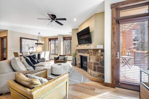 A spacious living room with a stone fireplace, a ceiling fan, and a large TV mounted on the wall. - Downtown condo with pool, hot tubs & gym - walk to lifts, near everything (Breckenridge)