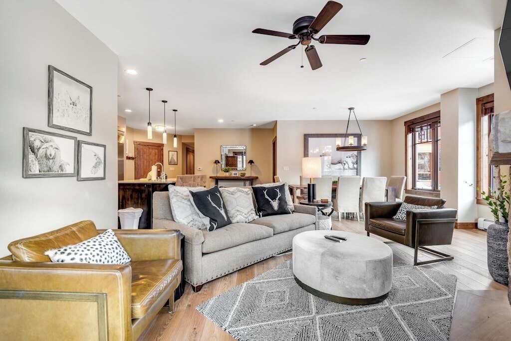 A living room with a sofa, armchair, ottoman, ceiling fan, and artwork on the walls.