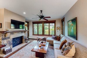 A living room with a stone fireplace, a ceiling fan, a large window, and a sofa. - Luxury condo near Peak 9 base with outdoor pool, hot tubs & gym (Breckenridge)