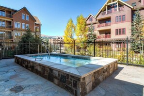 A swimming pool with a stone border, surrounded by a metal fence, with multi-story buildings in the background. - Luxury condo near Peak 9 base with outdoor pool, hot tubs & gym (Breckenridge)