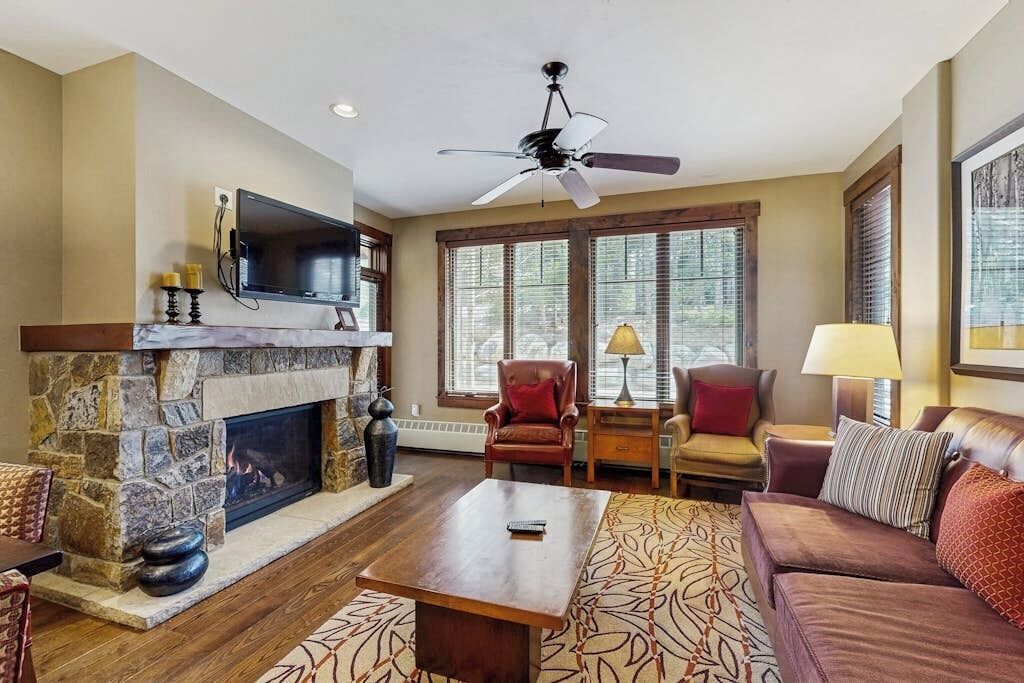 A living room with a stone fireplace, a ceiling fan, and a large window with blinds.