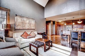 A spacious living area with a large sofa, a coffee table, and a dining table with chairs. There is a kitchen visible in the background. - Bright, luxury condo with pool, hot tubs & gym - walk to lifts & downtown (Breckenridge)