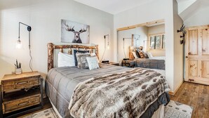 Een slaapkamer met een groot bed, een houten nachtkastje, een spiegel en een hertenkunstwerk aan de muur.