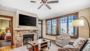 A living room with a stone fireplace, a flat-screen TV, a ceiling fan, and large windows. - Walk-to-lift condo with beautiful view, outdoor pool, hot tubs & fitness center (Breckenridge)