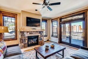 A living room with a stone fireplace, a flat-screen TV, and a ceiling fan. - Light-filled corner condo with great view, pool & hot tubs - walk to lifts (Breckenridge)