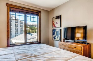 A hotel room with a large bed, a wooden dresser, a flat-screen TV, and a window with a mountain view. - Luxury condo with large balcony, great views, pool & hot tubs - walk to lifts (Breckenridge)