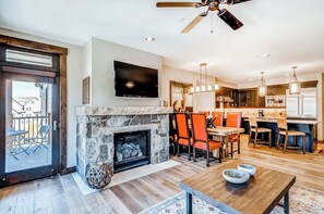 A modern living room with a stone fireplace, a flat-screen TV, and a dining area with orange chairs. - Luxury condo with large balcony, great views, pool & hot tubs - walk to lifts (Breckenridge)