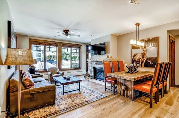 A living room with a sofa, coffee table, fireplace, dining table with chairs, and a TV mounted on the wall. - Luxury condo with large balcony, great views, pool & hot tubs - walk to lifts (Breckenridge)