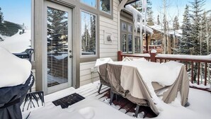 Une terrasse enneigée avec une table et des chaises, un espace extérieur abrité avec des sièges, et un bâtiment doté de grandes fenêtres et d’une porte.