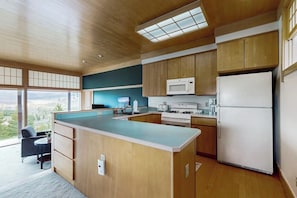 A modern kitchen with wooden cabinets, a central island, a microwave, a refrigerator, and a view through large windows. - Cozy home with full kitchen, private patio & mountain views (Grand Lake)