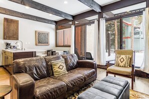 Ein Wohnzimmer mit Ledersofa, einem Holztisch und einem Sessel mit Kissen. Es gibt eine Küchenzeile mit Arbeitsplatte und Schränken, sowie ein großes Fenster mit Jalousien.