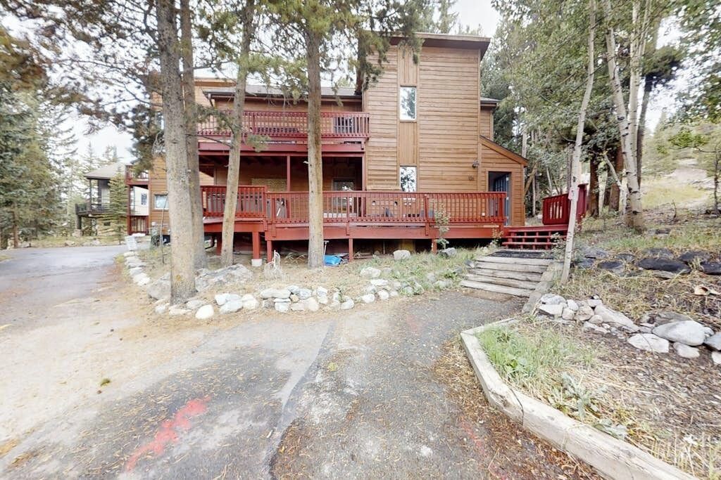 A two-story wooden cabin with a balcony, surrounded by trees and a gravel path.