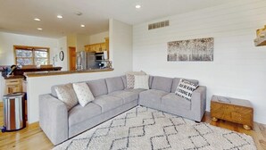 A living room with a grey sectional sofa, a ceiling fan, and a kitchen in the background. - Stunning house w/ mountain views, free WiFi, outdoor activities (Granby)