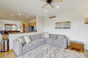 Ein Wohnzimmer mit einem grauen Ecksofa, einem Deckenventilator und einer Küche im Hintergrund.